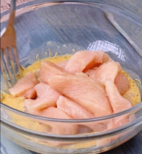 raw chicken strips being mixed with beaten eggs in a glass bowl