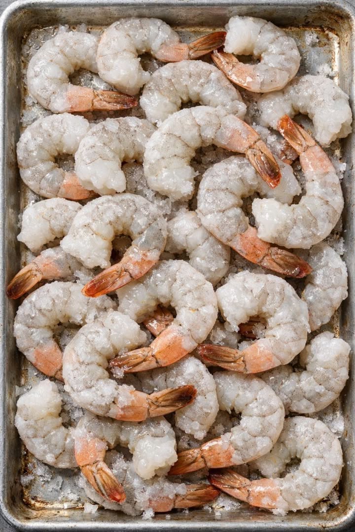 Frozen raw peeled shrimp with tails arranged on a metal tray before making garlic butter shrimp.